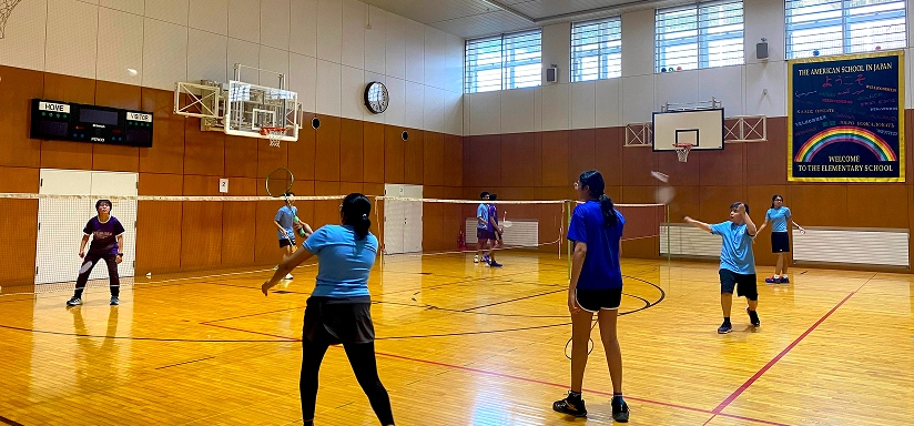 Students play badminton in a school gym, engaging in a doubles match with focus and energy.