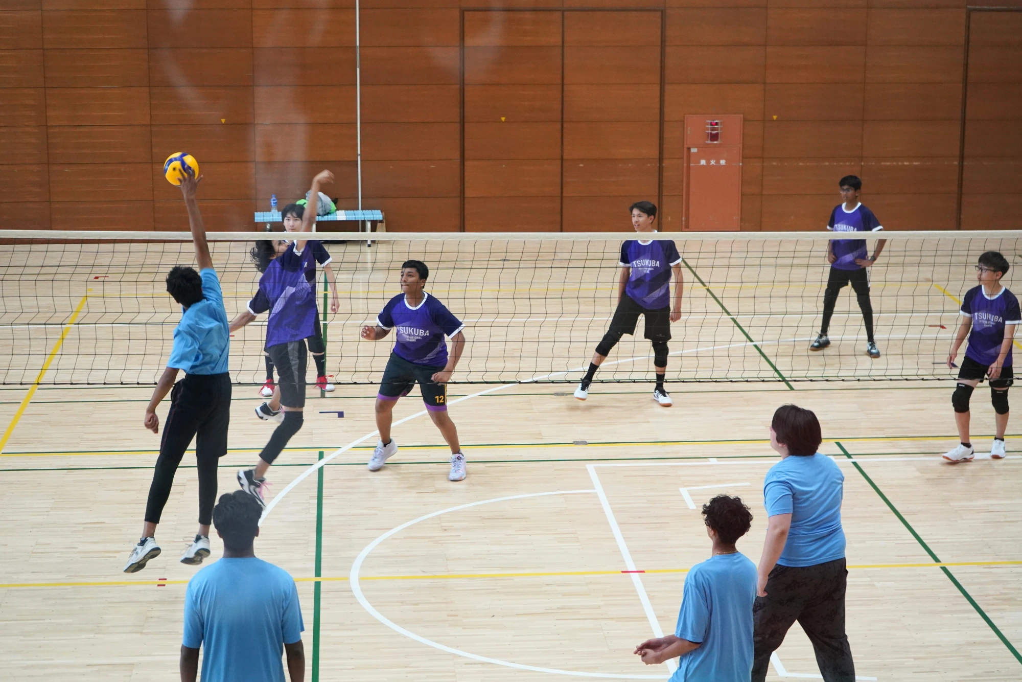 Indoor volleyball game with players in blue and purple jerseys, player in blue spiking the ball over the net.