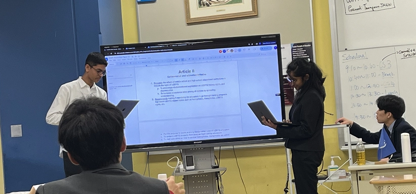 Students engage in a classroom presentation and debate with laptops and a large screen displaying text.