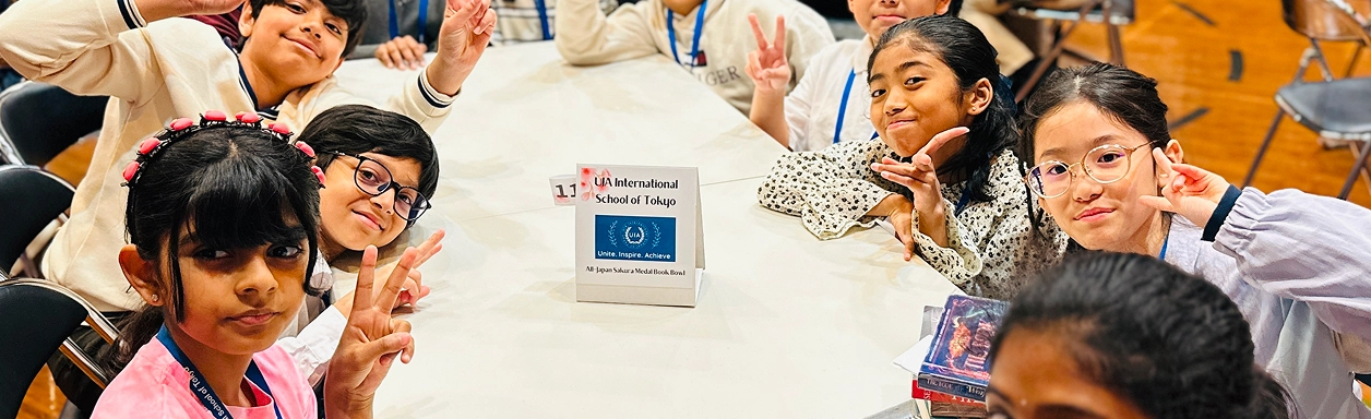 Children gathered around a table at UJA International School Tokyo, smiling and holding up peace signs.