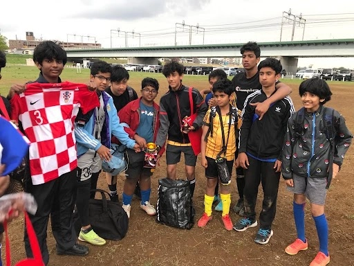 Youth soccer team gathers on muddy field with jerseys and gear bags; overcast sky and bridge in background.