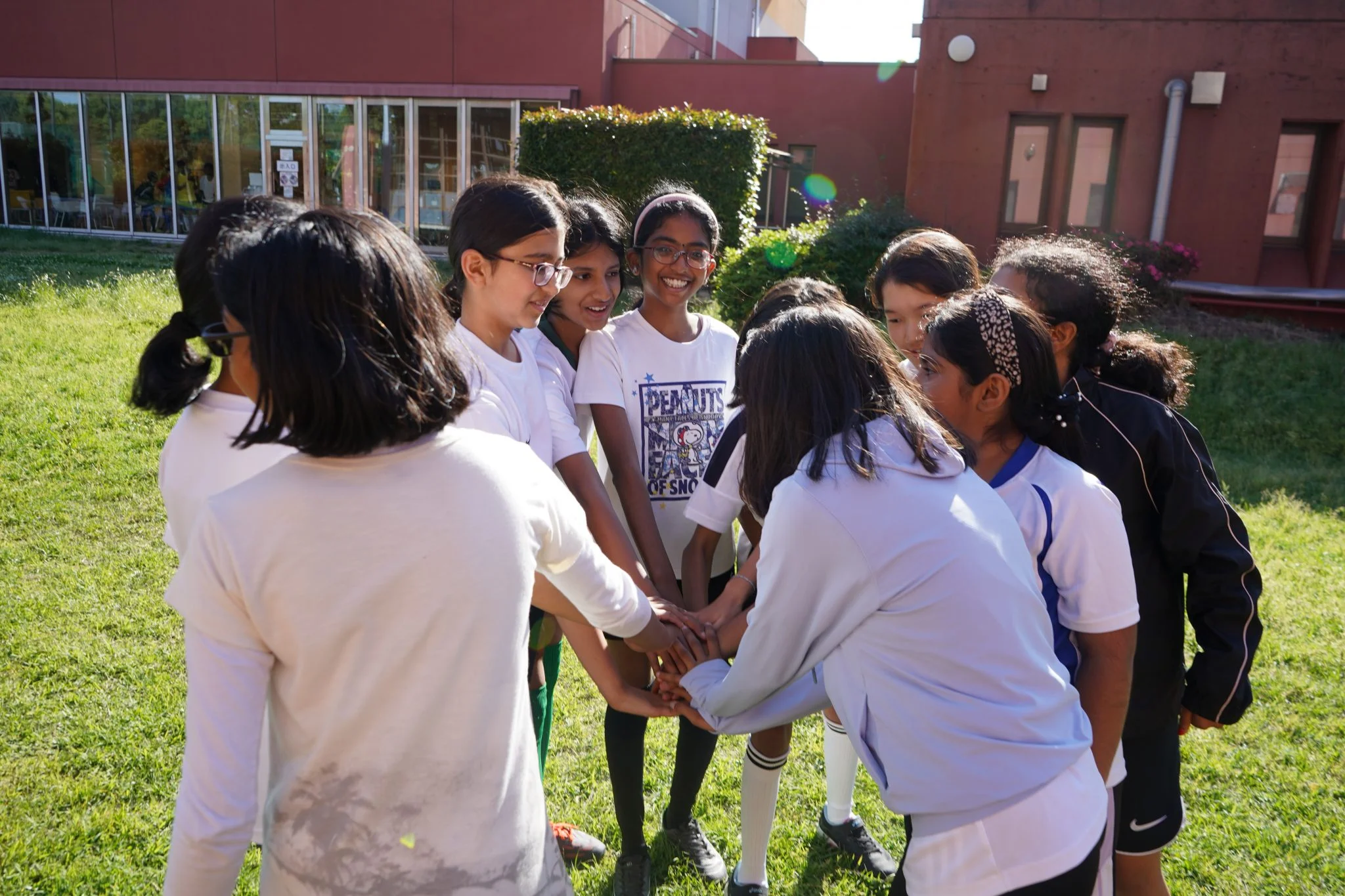 Group of girls in a team huddle outdoors, united hands in center, building teamwork and friendship in a school setting.