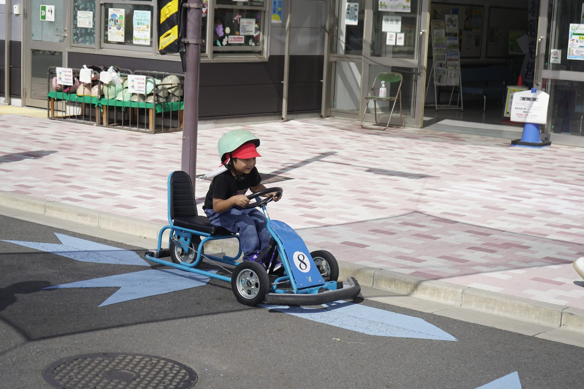 Child driving a blue pedal kart on a sunny day, wearing a helmet and cap, outdoors near a building.
