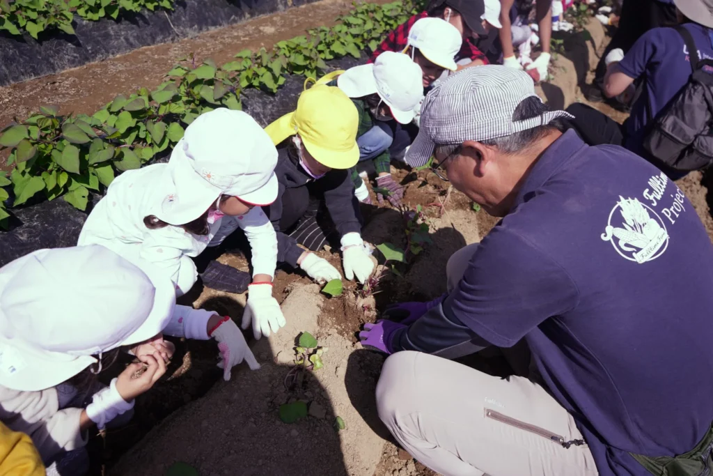 Children learning to plant in a garden with a teacher, wearing hats and gloves, engaging in hands-on agriculture.