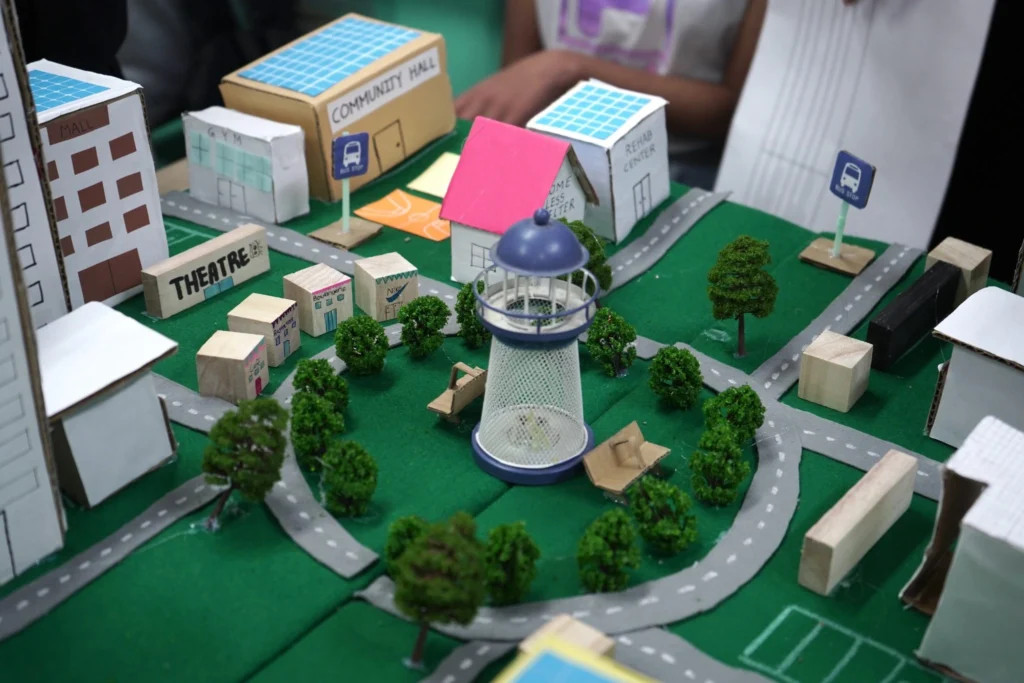Model of a green community with miniature buildings, roads, trees, and a central lighthouse, promoting sustainable living.
