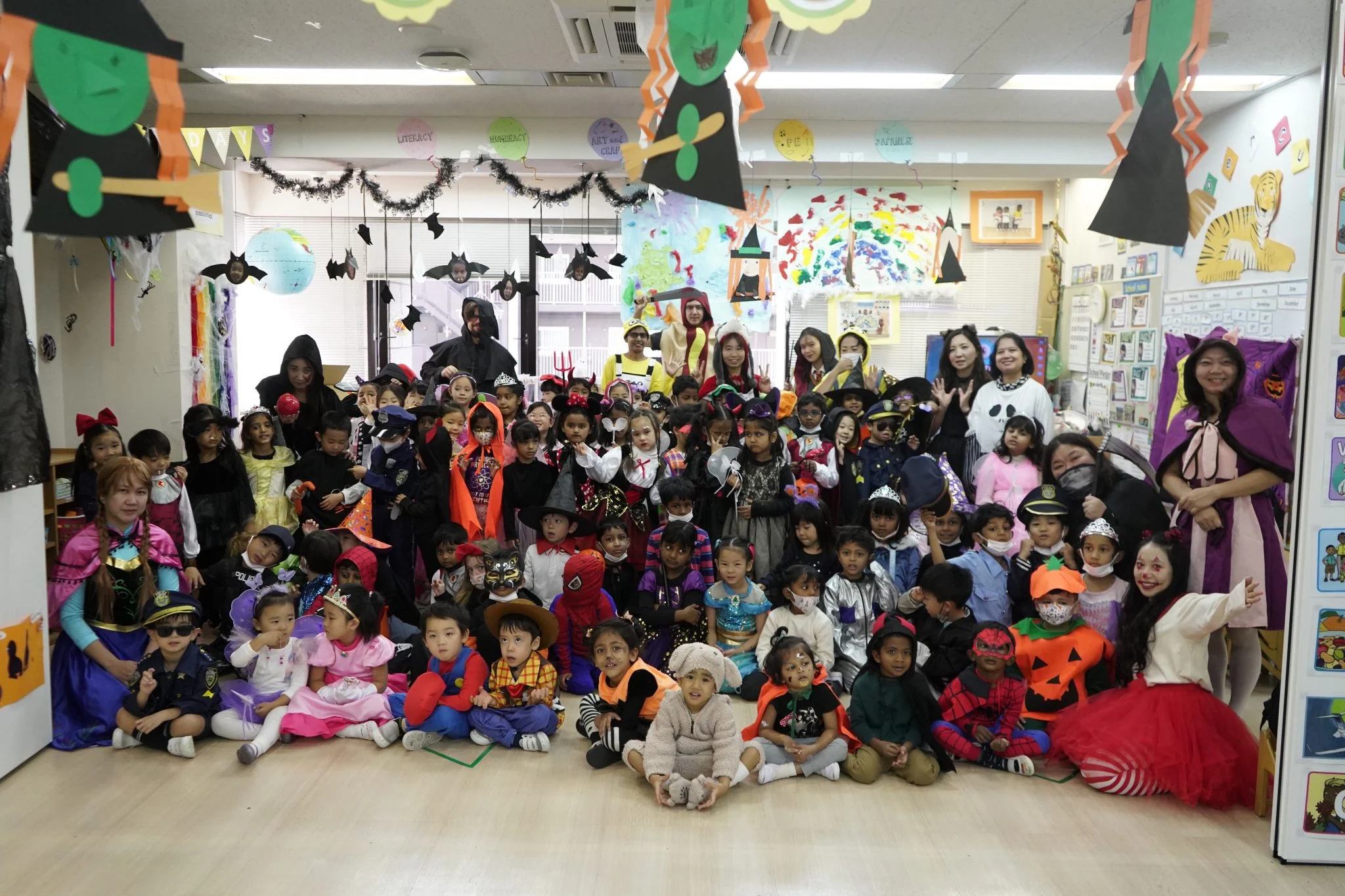 Children and teachers in colorful Halloween costumes gathered in a festive classroom setting.