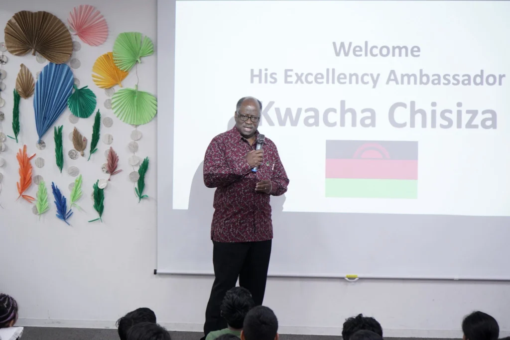 Man speaking at an event with colorful decorations, projected text with Welcome His Excellency Ambassador, flag in view.