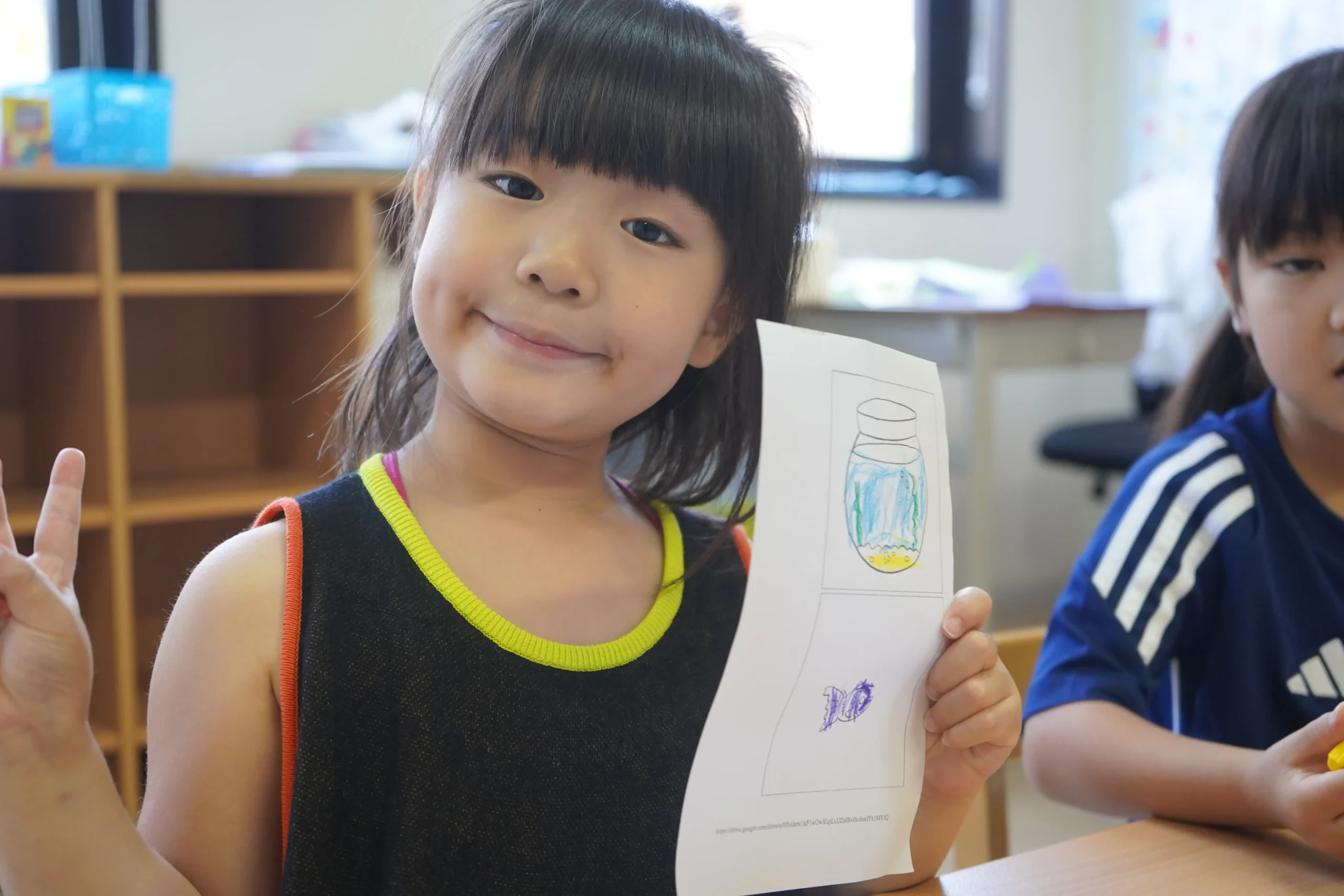 Child holding a colorful drawing, smiling and making a peace sign in a classroom setting.