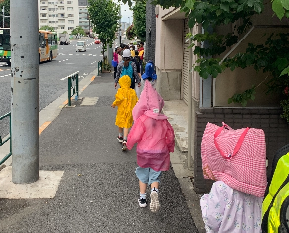 Children walking on a city sidewalk in colorful raincoats and backpacks, with traffic in the background.