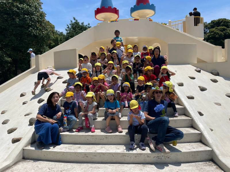 Group of children with yellow hats and teachers posing on a playground slide under a sunny blue sky.