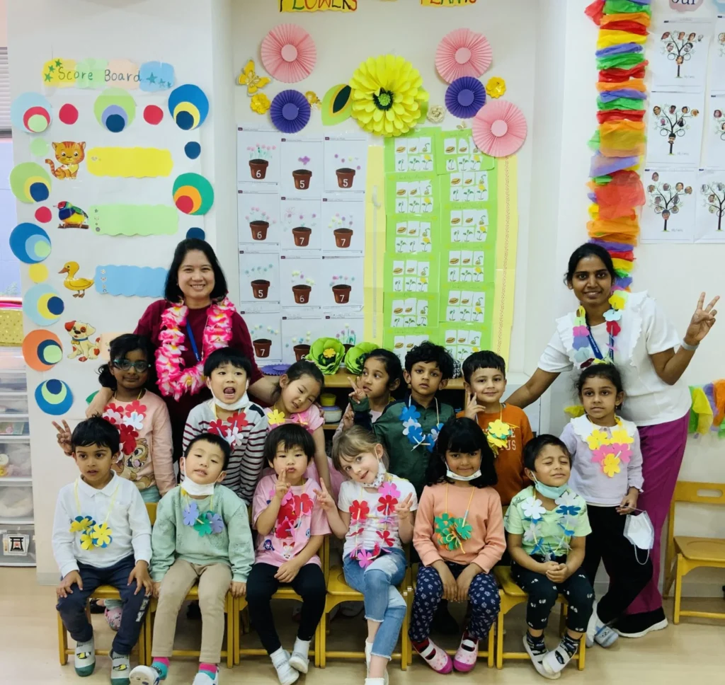 Preschool class group photo with teachers and children wearing colorful flower crafts, classroom decorations in the background.