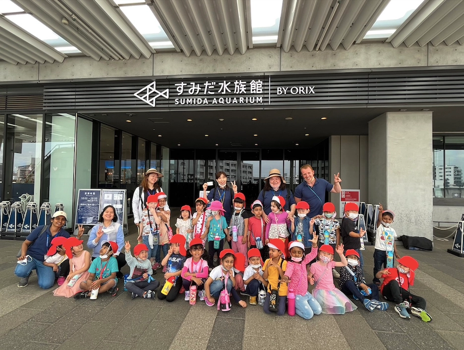 Group of children and teachers at the Sumida Aquarium entrance, smiling and wearing red hats.