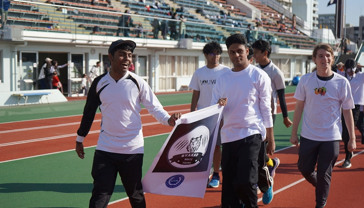 Group of young people walking on a track, holding a banner at a sports event, wearing white and black sports attire.