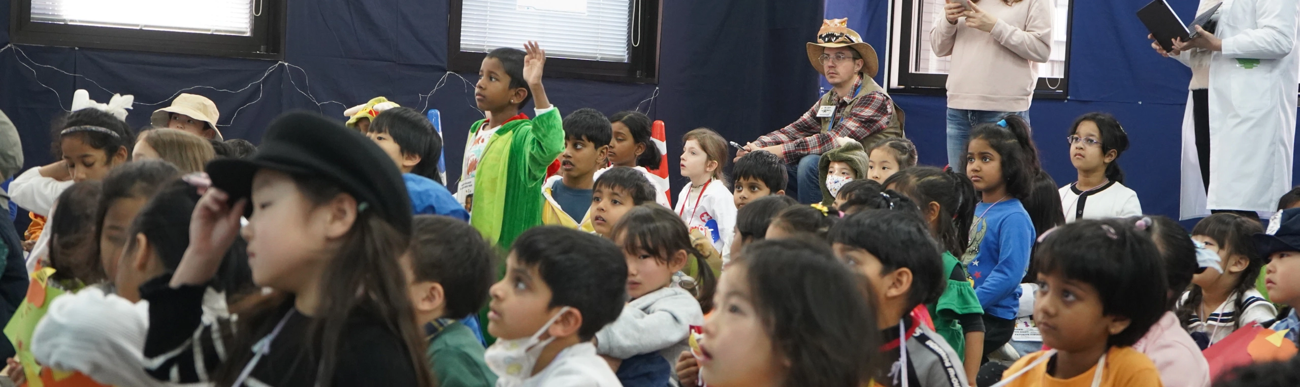 Children attentively watching a performance at a school event, one raising hand, diverse group in colorful clothing.