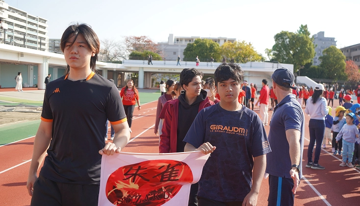 A group of young people walking on an athletic track during a sports event, holding a banner with red and black design.