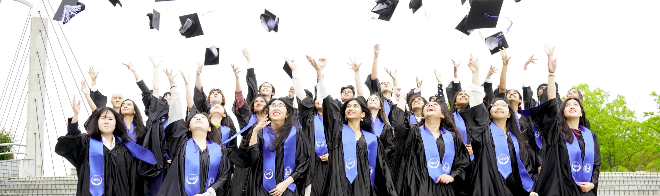 Graduates celebrating by tossing caps in the air, wearing gowns and blue sashes, outdoors on graduation day.