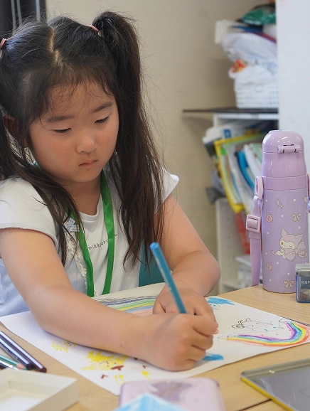 Young girl drawing with blue pencil at a table, surrounded by art supplies and a purple thermos.