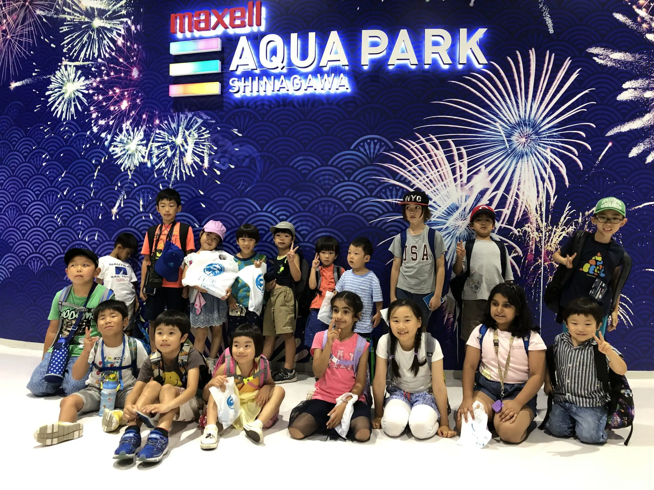 Group of children smiling at Maxell Aqua Park Shinagawa, with vibrant fireworks display in the background.