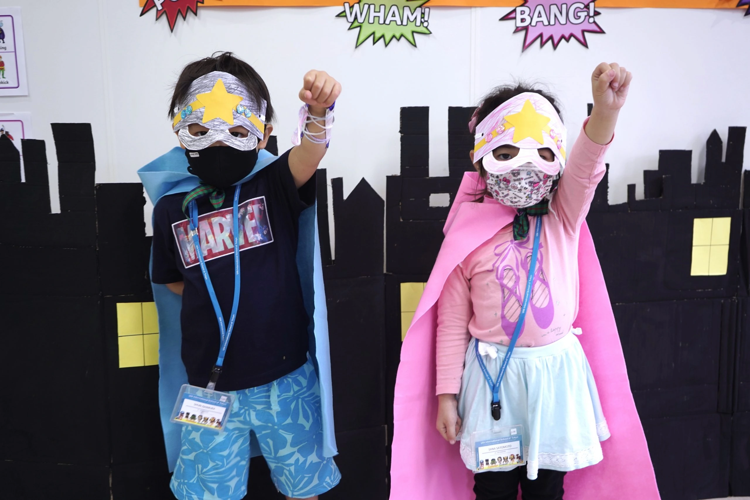 Children dressed as superheroes with colorful capes and masks, posing with fists up in a playful classroom setting.