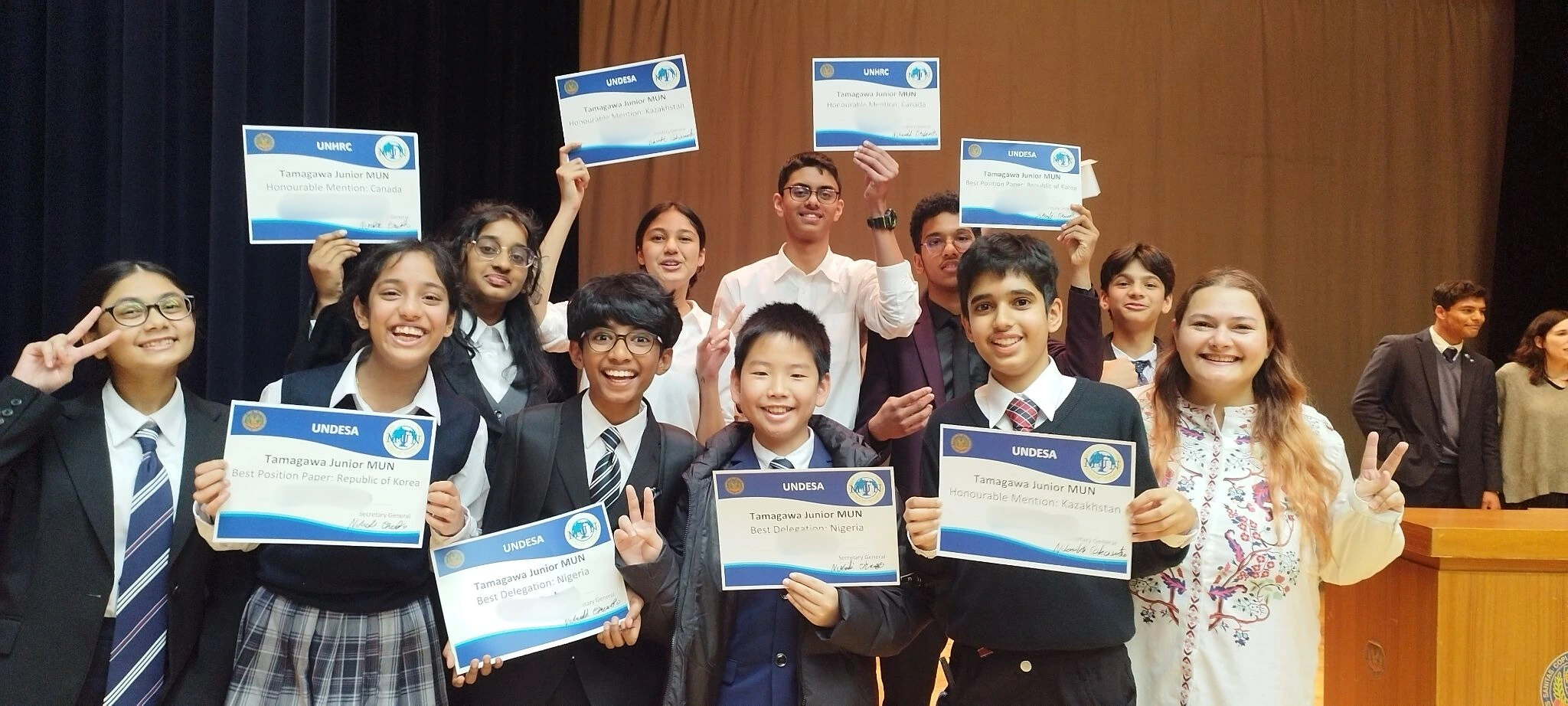 Students celebrate with awards at Tamagawa Junior MUN event, holding certificates and smiling on stage.
