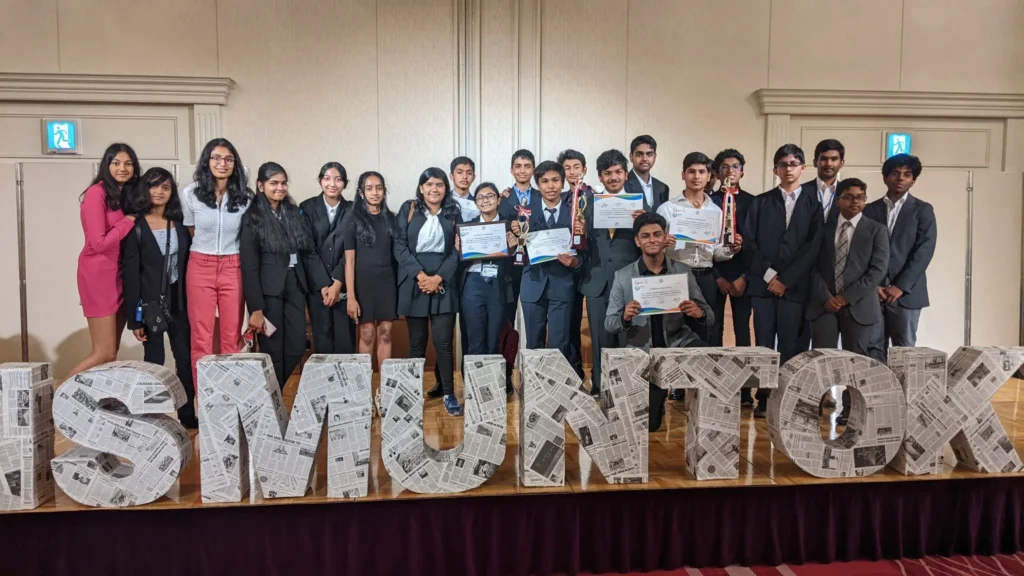 Group of students in formal attire holding certificates and trophies at an event, standing behind large ISMUN TOK letters.