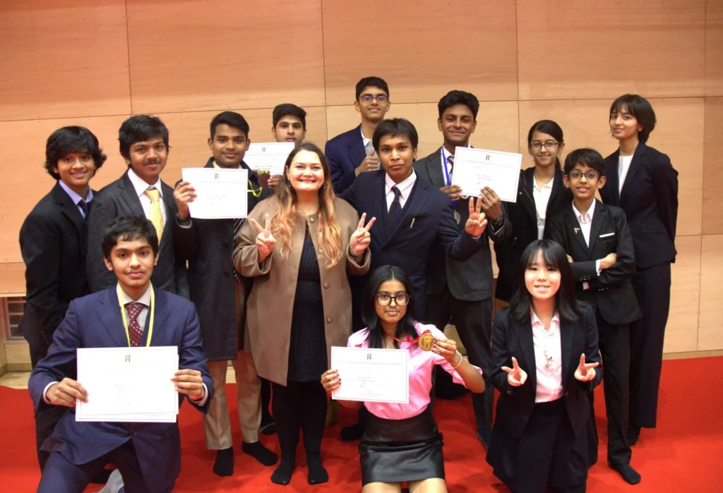 A group of students in formal attire pose with certificates and medals, celebrating their achievement on a red carpet.