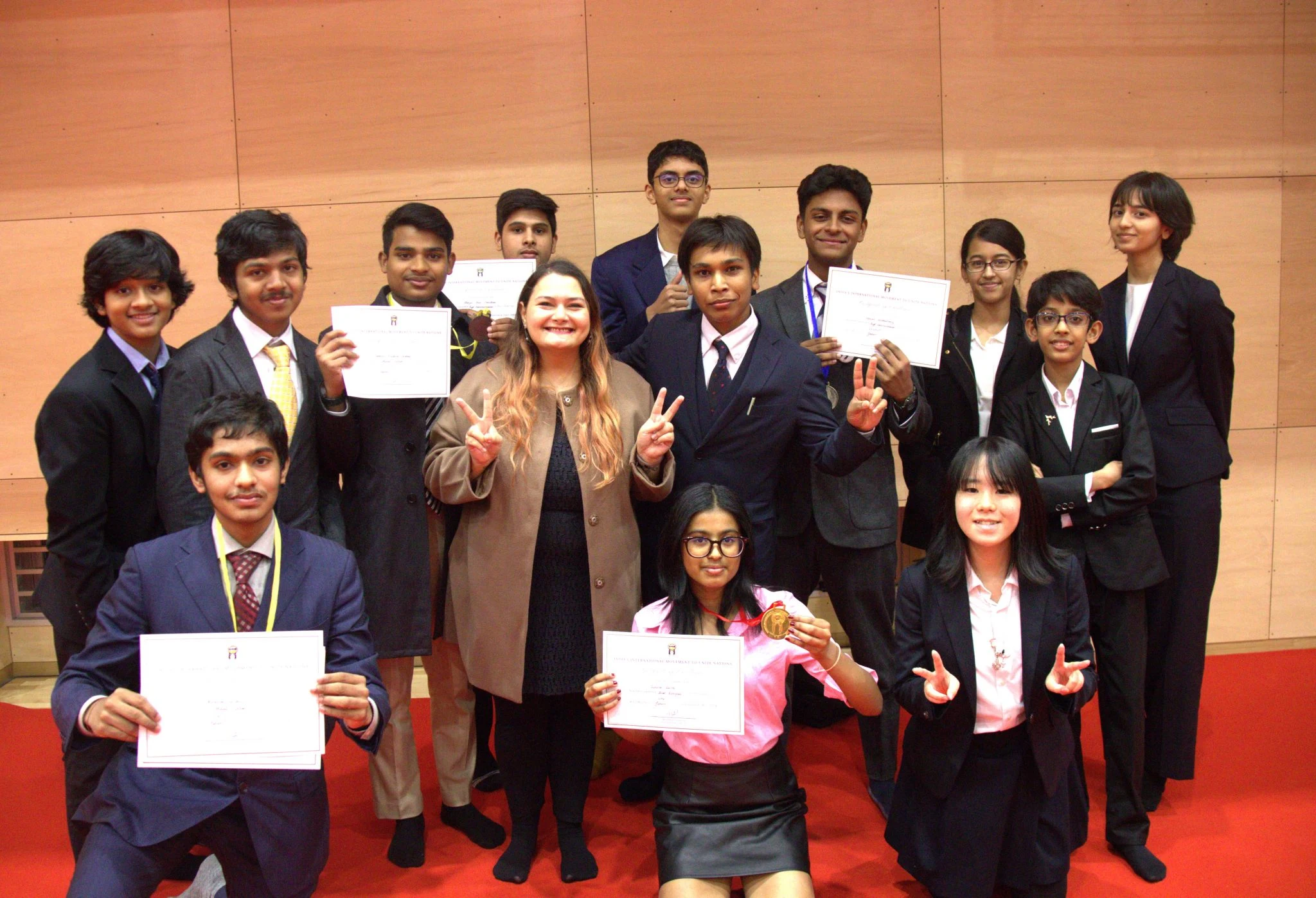 A group of students in formal attire pose with certificates and medals, celebrating their achievement on a red carpet.