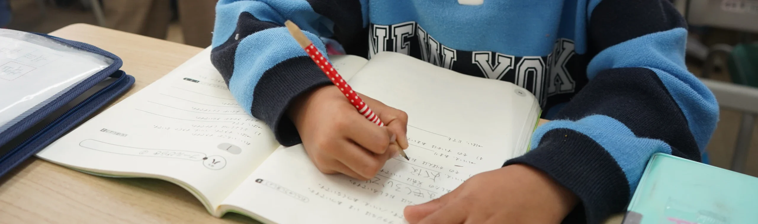 Child writing in a notebook with a red pencil in a classroom setting.