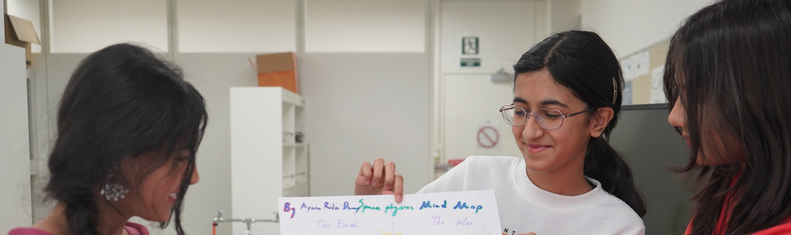 Three students presenting a colorful space-themed mind map in a classroom setting.