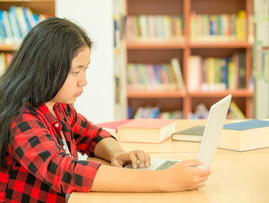 Student focused on laptop in a library, surrounded by books, wearing a red plaid shirt.