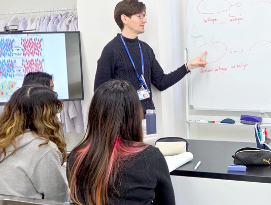 Teacher explaining scientific concepts to engaged students in a classroom with a whiteboard and presentation screen.