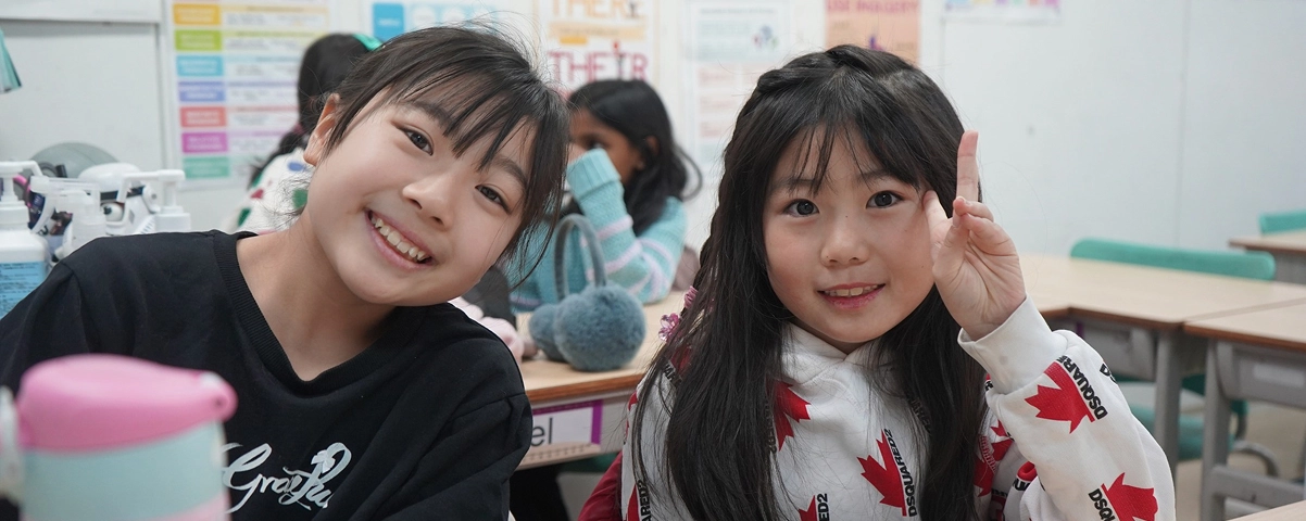 Two smiling girls in a classroom, one making a peace sign, seated at a desk with educational posters in the background.