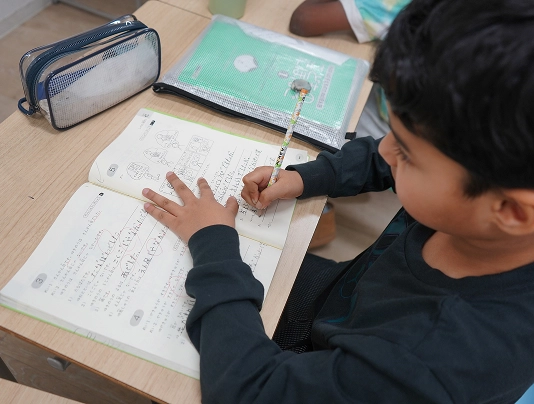 Child writing in notebook at a desk with pencil case nearby, focused on homework in a classroom setting.