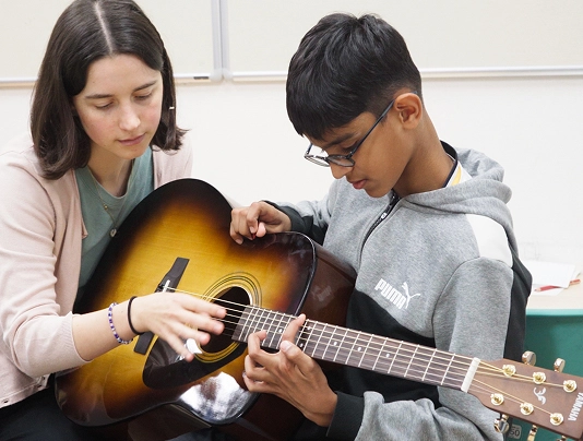 Guitar lesson with a tutor guiding a young student on acoustic guitar playing in a classroom setting.