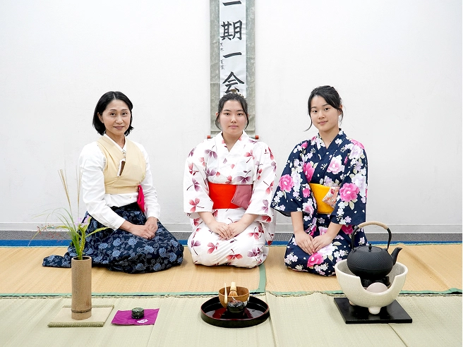 Three women in traditional kimonos pose for a Japanese tea ceremony, seated with tea set on tatami mats.