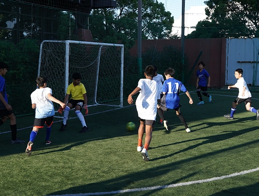 Children playing a competitive soccer match on a green field near the goal under a sunny sky.