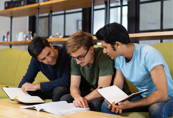 Three young men studying together, sitting on a couch with open books and notebooks, collaborating on a project.