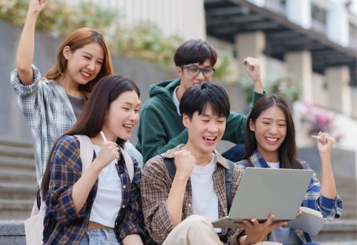 Five excited students celebrating success while looking at a laptop on campus steps.