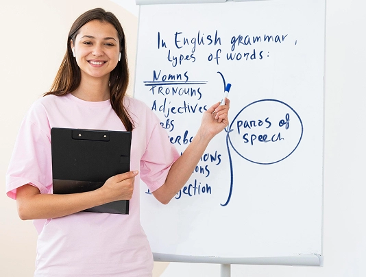 Smiling teacher explaining English grammar types on whiteboard in classroom, holding clipboard.