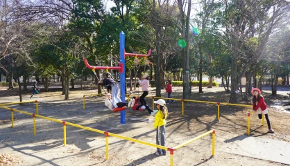 Children playing on a colorful playground merry-go-round in a sunny park surrounded by trees.