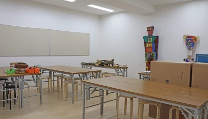 Empty classroom with art supplies and stacked cardboard boxes, featuring tables, chairs, and bright lighting.