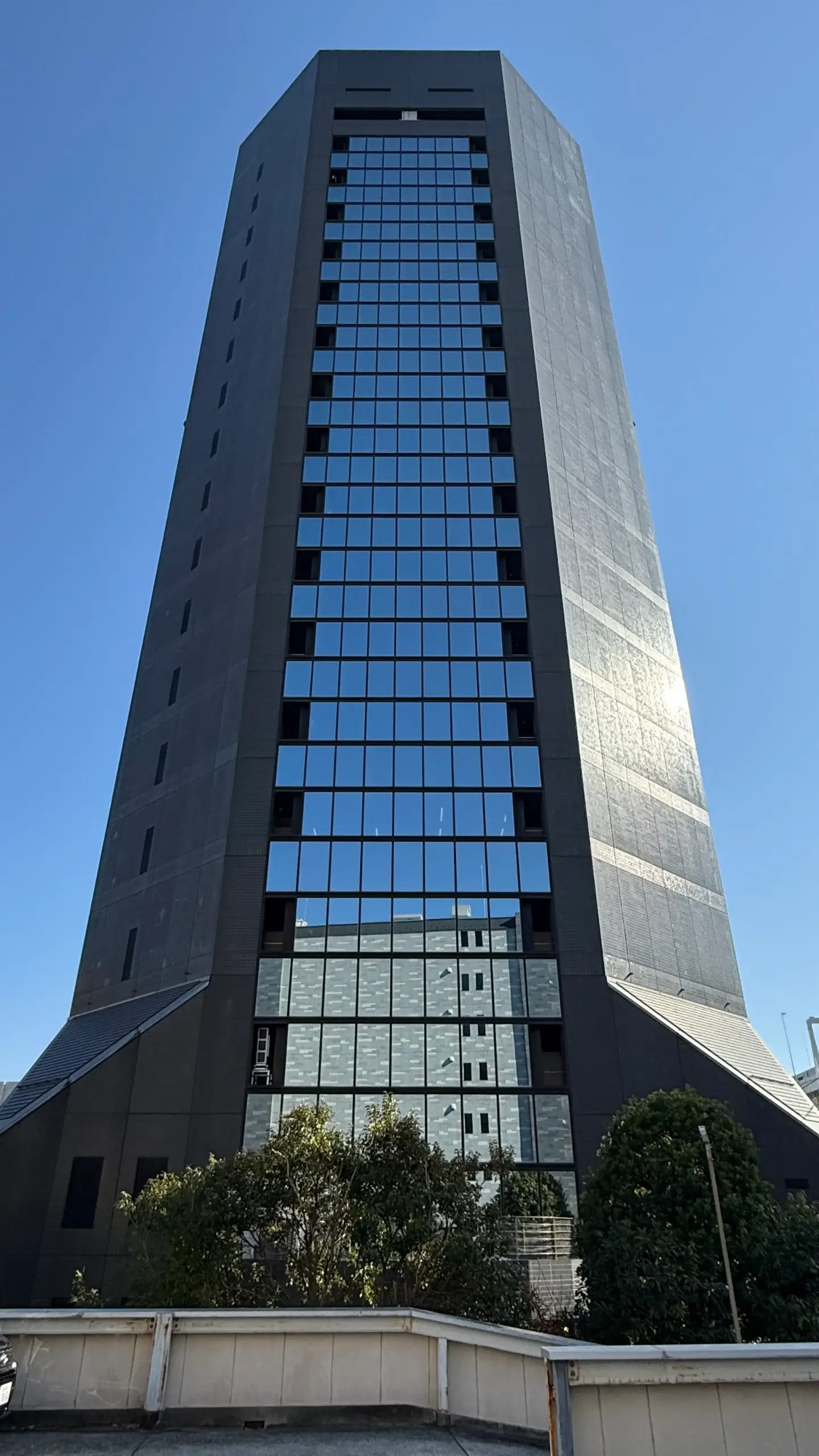 Tall modern office building with glass windows reflecting blue sky, surrounded by greenery.