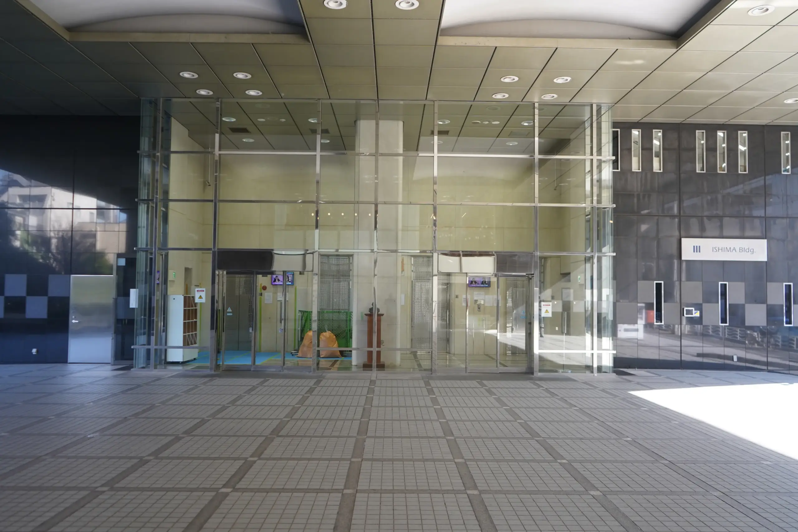 Modern glass entrance of Ishima Building with tiled flooring and overhead lighting during daylight.